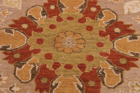 A close-up of a traditional patterned rug featuring a central floral medallion in muted yellow-green, surrounded by rust red, ochre orange, and brown motifs on a beige background.